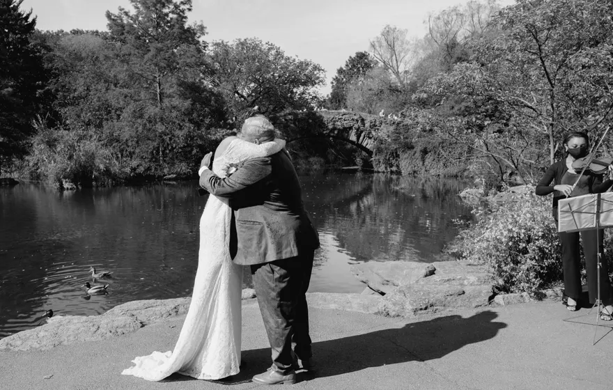 Black and white photo of a couple in a long embrace at the water's edge in Central Park, Gapstow Bridge behind them and a musician playing to one side