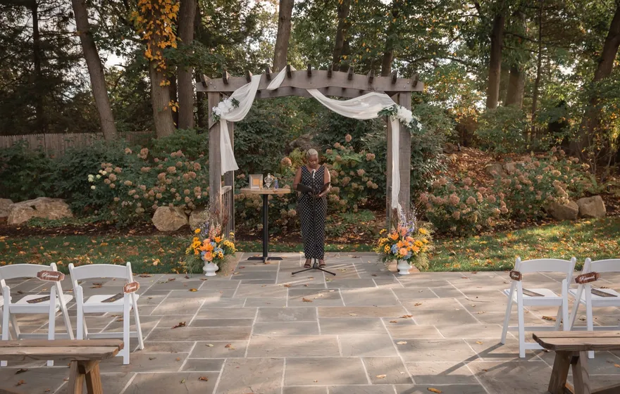 A garden ceremony site with a wooden arbor draped in white fabric, potted flowers at its base, the officiant standing at a microphone and white chairs on the stone patio