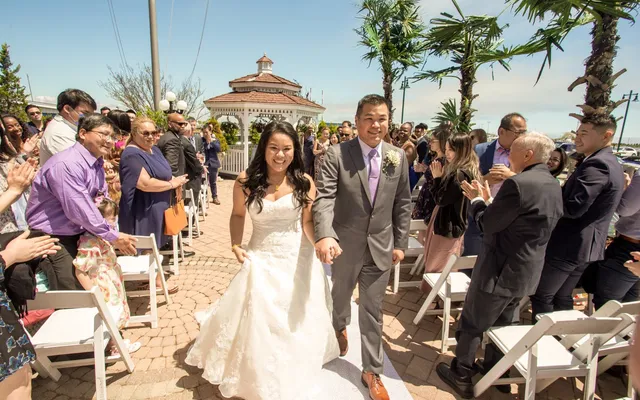 A bride and groom walk up the aisle between applauding guests with a white gazebo behind them