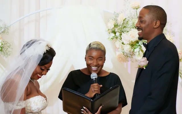 An officiant smiles as she speaks into a microphone with her ceremony binder open, the bride and groom laughing on either side