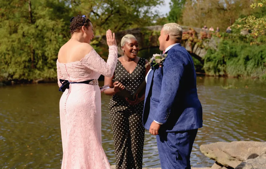 An officiant in a polka-dot jumpsuit raises one hand as she speaks to a couple renewing their vows beside the water in Central Park