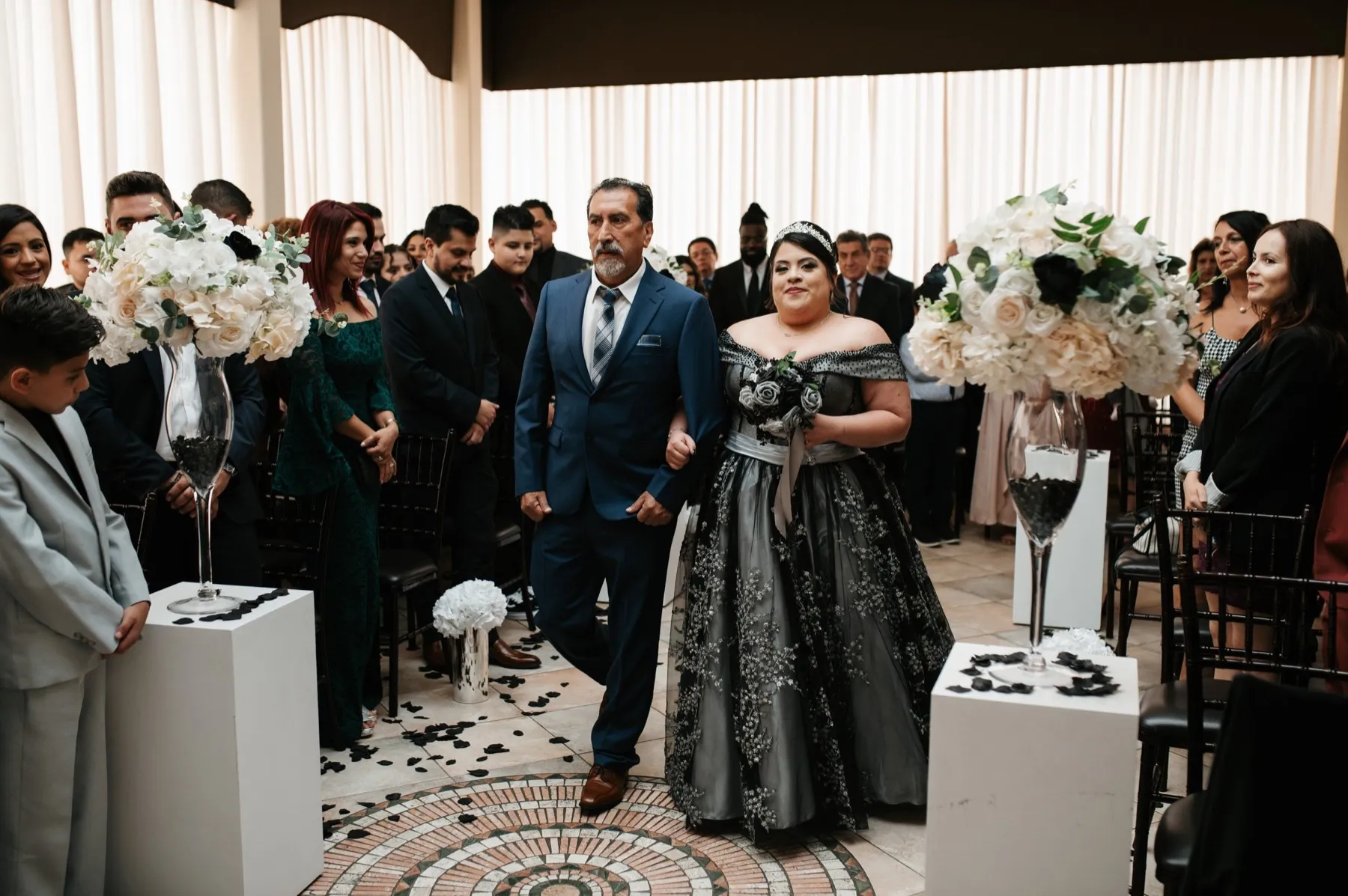 A bride in a black and silver gown walks down the aisle on her escort's arm as guests stand and watch