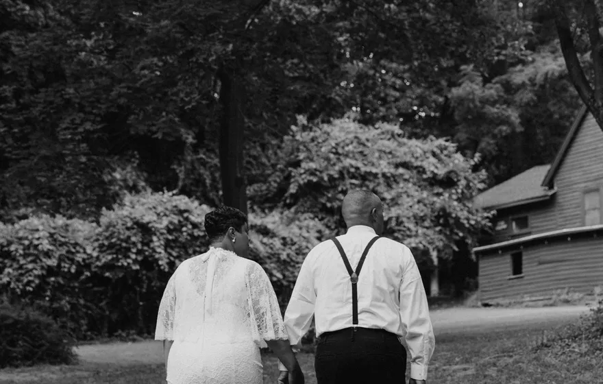 Black and white photo of a couple walking away hand in hand across a lawn toward the trees