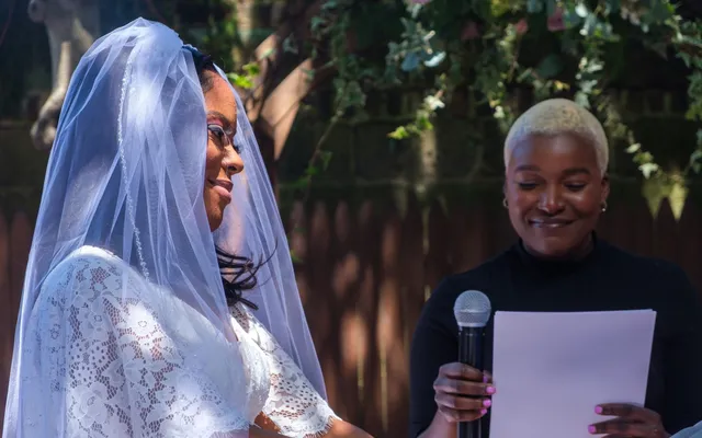 An officiant reads from a sheet of paper at a microphone while a bride in a lace dress and veil listens