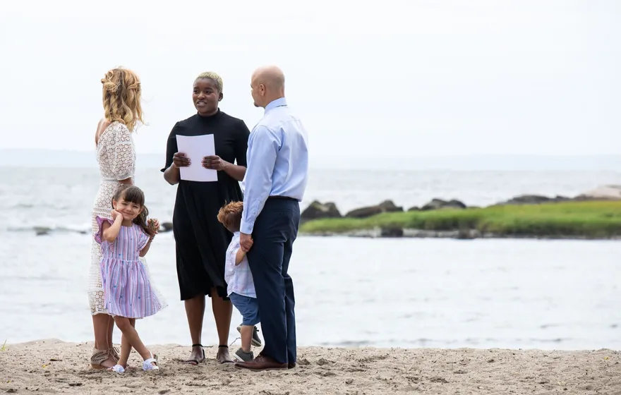 An officiant reads from a sheet of paper on a beach while the couple stands with their two young children, one girl twirling in a lilac dress