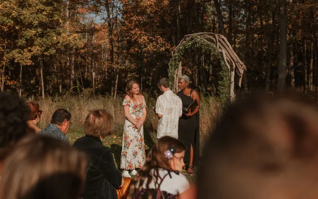 A small outdoor ceremony seen from among the seated guests, the couple and officiant standing before a branch arch in autumn woods