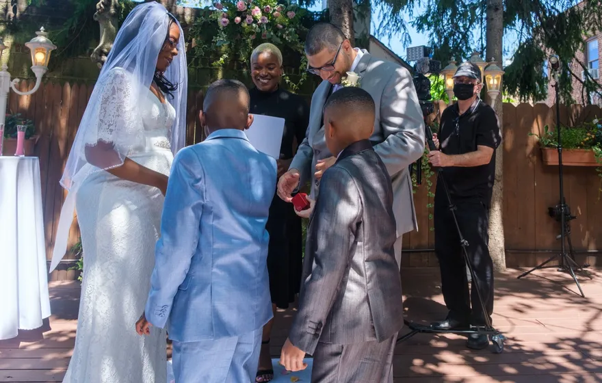 Two boys in light and dark gray suits stand at the front with a bride in a veil and a groom holding a red ring box, the officiant between them