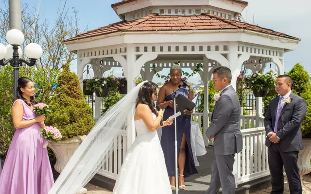 A bride reads from a page to her groom in front of a white gazebo while the officiant holds the microphone