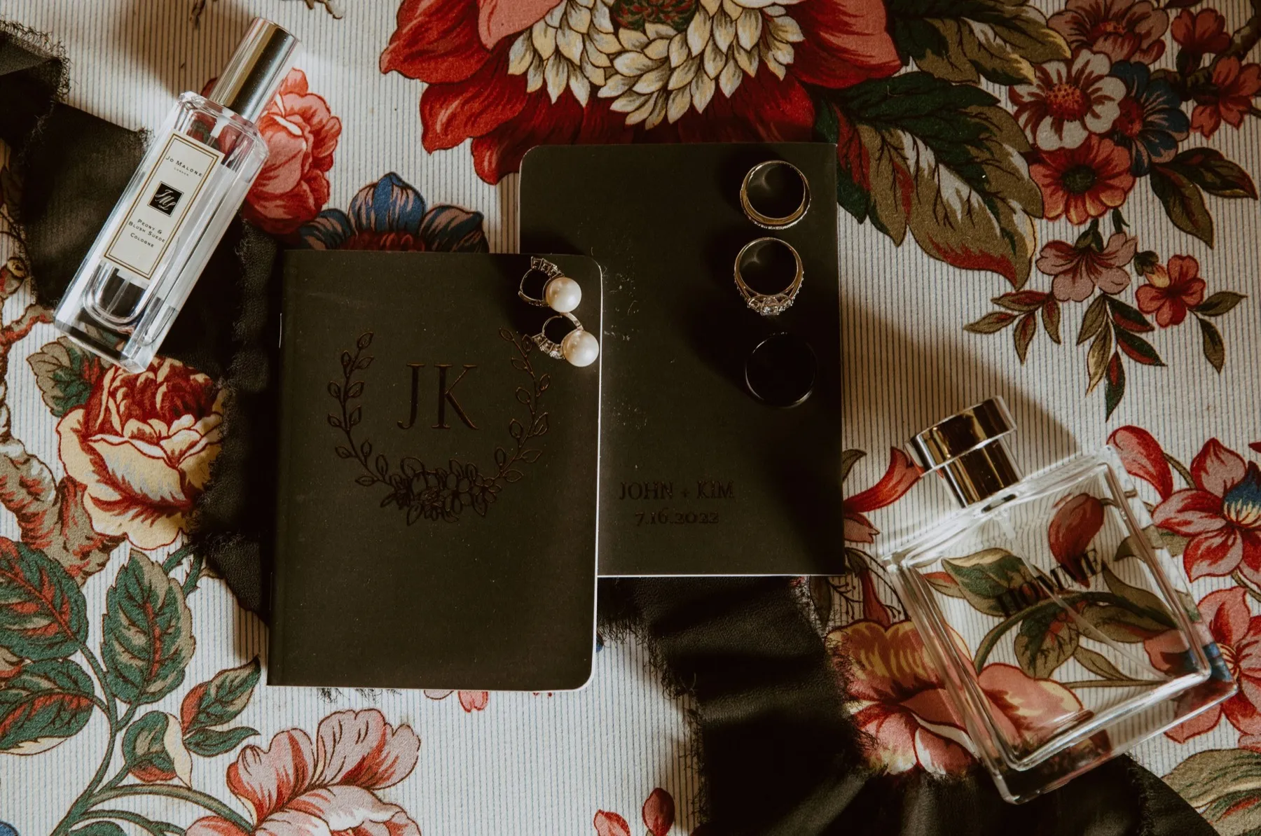 Two black monogrammed vow books laid on floral fabric with rings, pearl earrings and perfume bottles