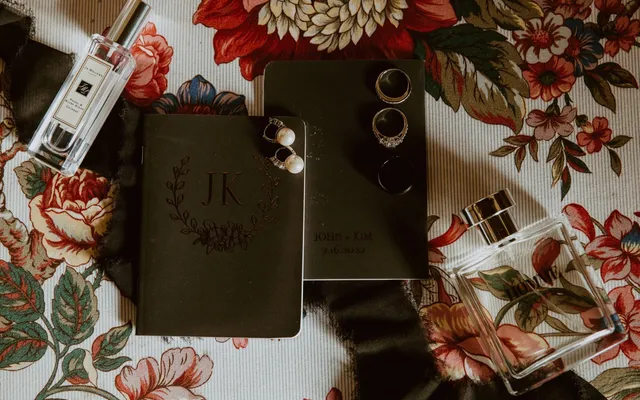 Two black monogrammed vow books laid on floral fabric with rings, pearl earrings and perfume bottles