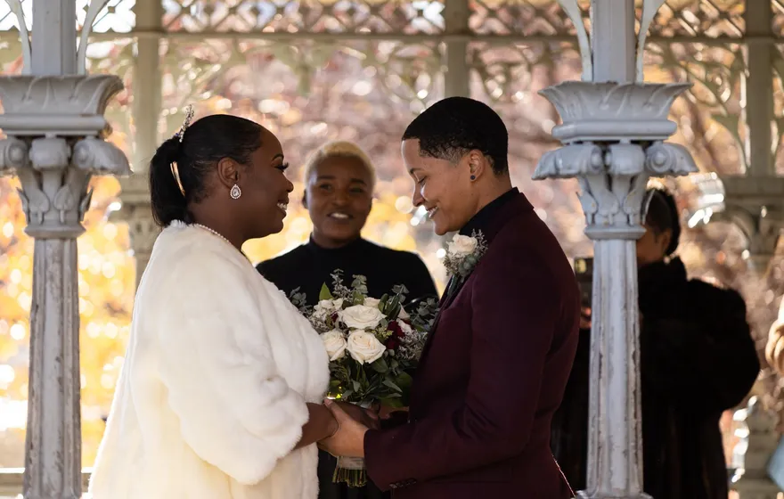 Two partners, one in a white fur wrap and one in a burgundy suit, hold hands and smile under the ironwork of the Ladies' Pavilion while the officiant stands behind them