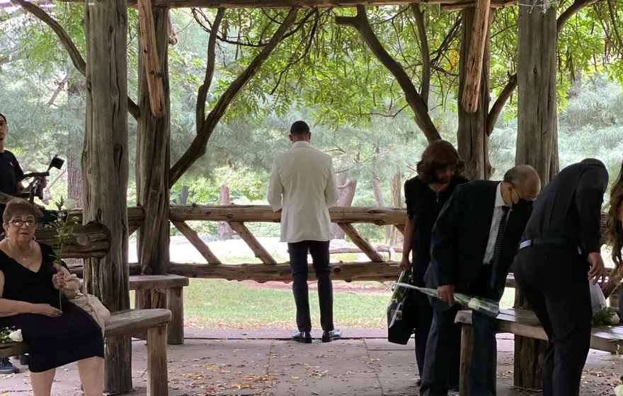 Guests standing and sitting on built-in benches under the rustic wooden Cop Cot shelter in Central Park, fallen leaves on the stone floor