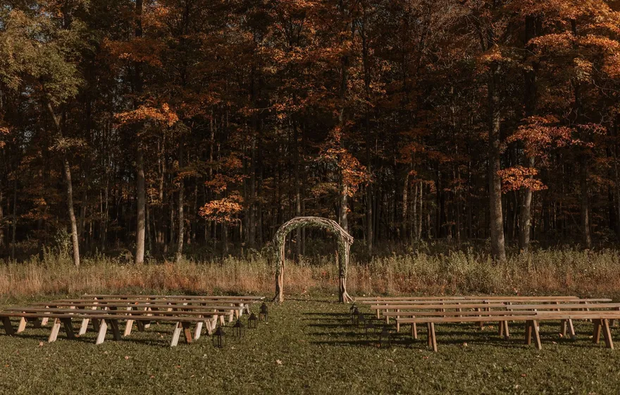 Rows of wooden benches facing a branch arch at the edge of autumn woods, set up for an outdoor ceremony