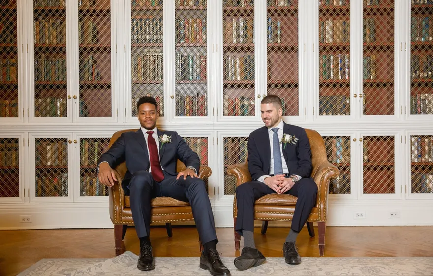 Two grooms in navy suits sit side by side in leather armchairs in front of tall latticed bookcases