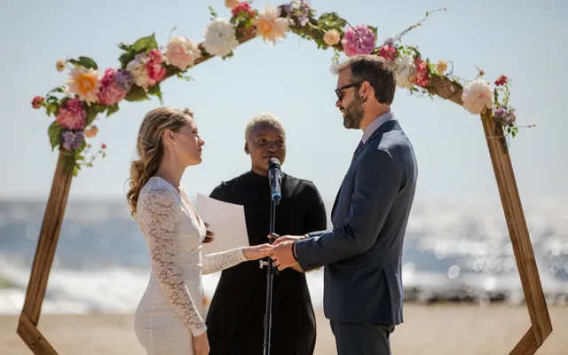 A couple holds hands under a flower-covered wooden arch on a beach while the officiant stands between them at a microphone