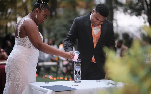 A bride and groom pour colored sand into an hourglass on a white-clothed table outdoors