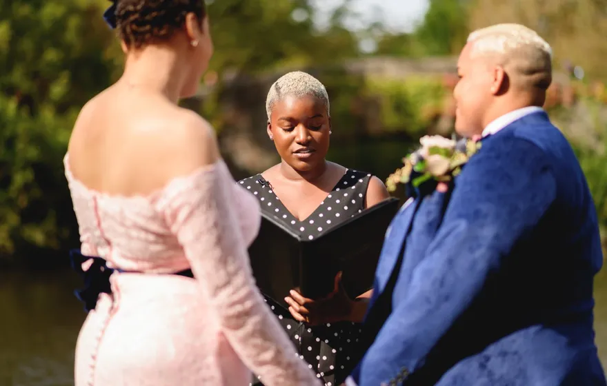 The officiant reads from an open black binder while a couple in a pink lace gown and a blue suit face each other by the water