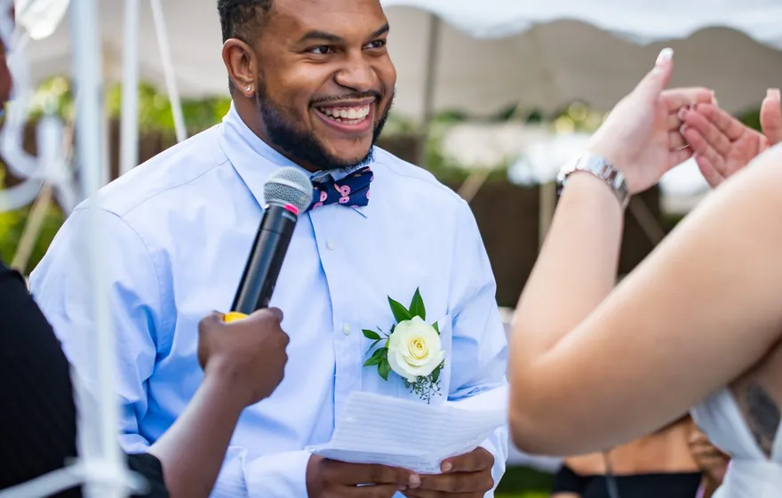 A groom in a light blue shirt and bow tie grins as he holds a folded card of vows and a microphone at an outdoor ceremony