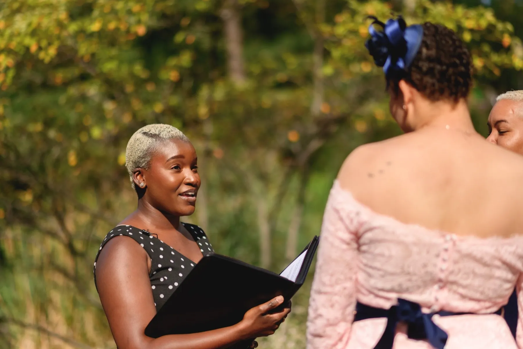 An officiant with an open ceremony binder speaks to a couple during an outdoor vow renewal