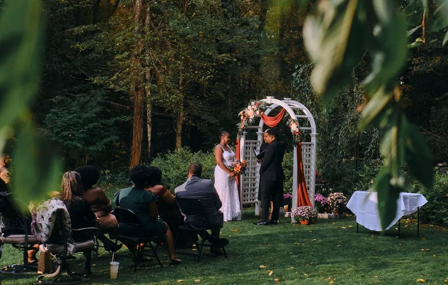 An outdoor ceremony seen through leaves, guests seated on the lawn facing a white arch at the edge of tall trees