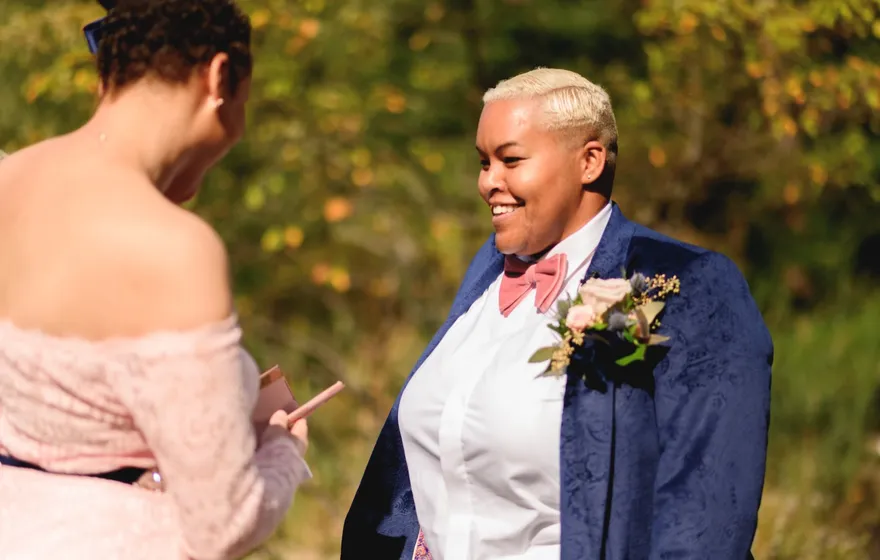 One partner reads from a small card while the other, in a navy jacket and pink bow tie, smiles back at her