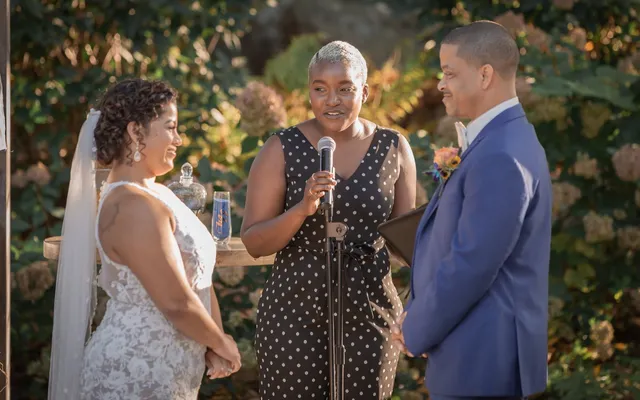 An officiant in a polka-dot jumpsuit speaks into a microphone between a bride and groom in a garden