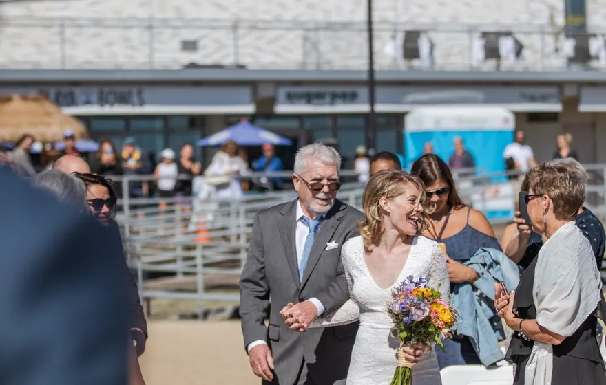 A bride carrying a bouquet walks down a sandy beach aisle on an older man's arm, guests standing beside rows of white chairs