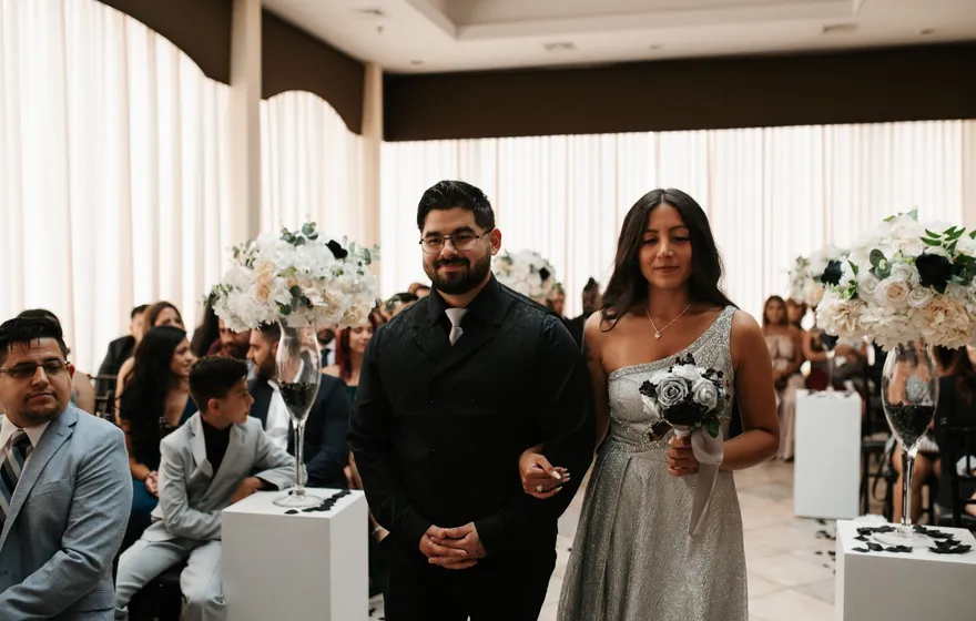 A groomsman in a black suit and a bridesmaid in a silver gown walk arm in arm down a white aisle between standing guests