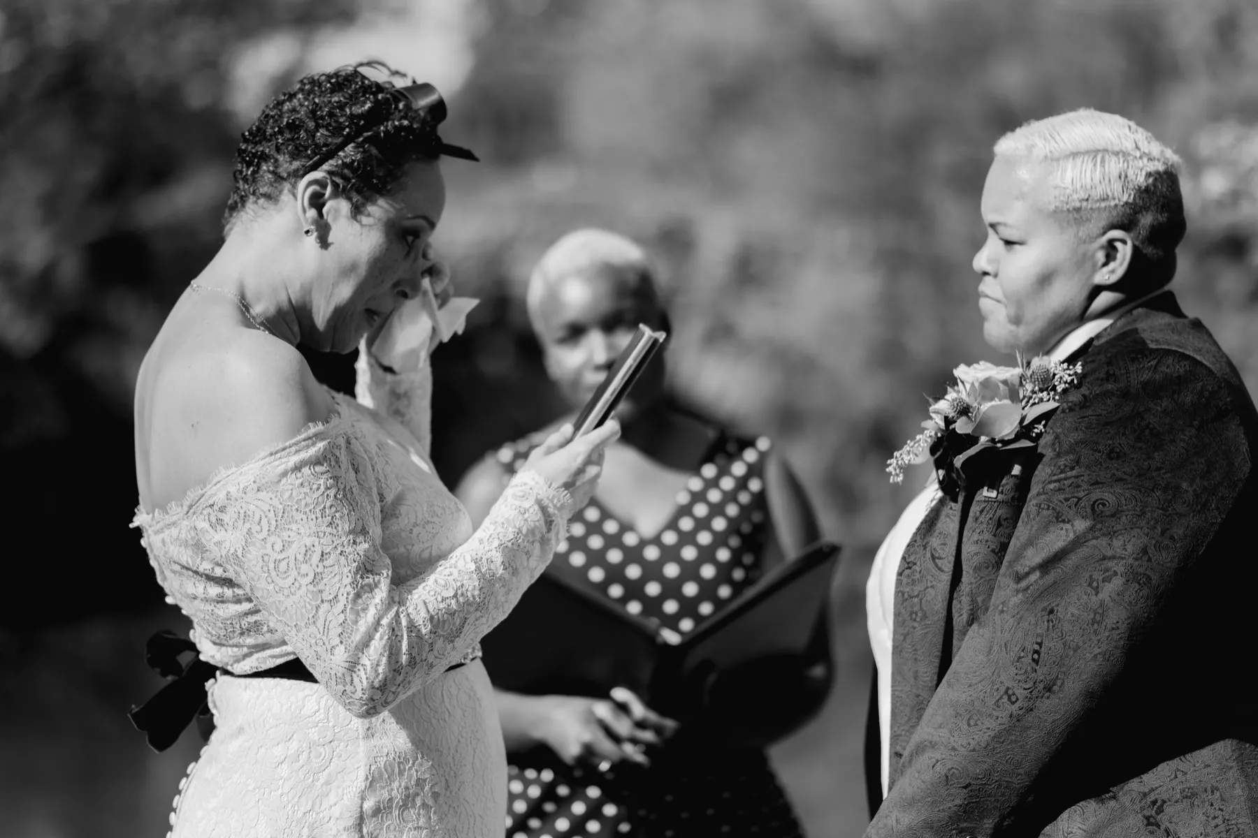 Black and white photo of a woman wiping away a tear as she reads her vows from her phone, her spouse and the officiant looking on
