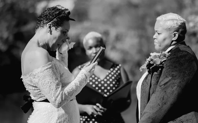 Black and white photo of a woman wiping away a tear as she reads her vows from her phone, her spouse and the officiant looking on