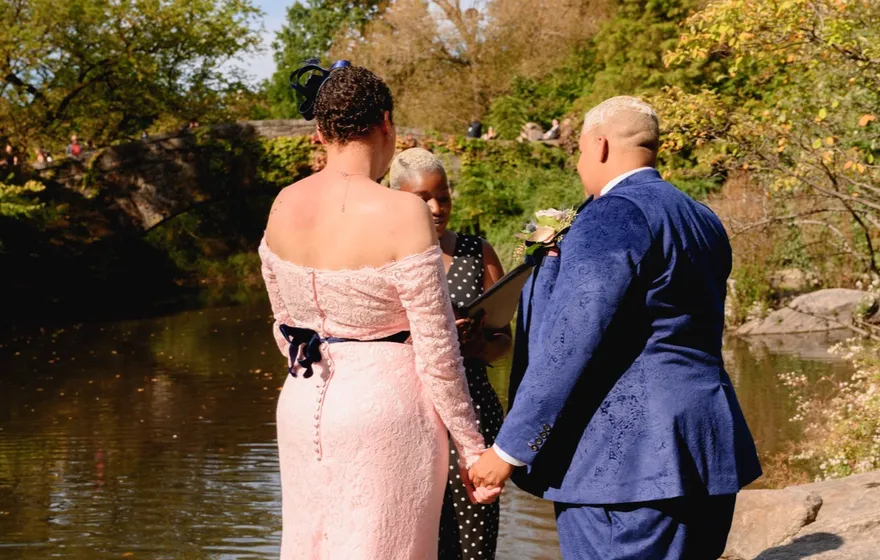 A couple in a pink lace gown and a blue suit hold hands on the rocks at the edge of the lake in Central Park, the officiant just behind them