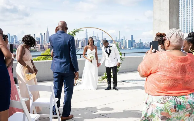 A guest stands in the aisle holding up a phone to film a rooftop wedding ceremony, the Manhattan skyline behind the couple