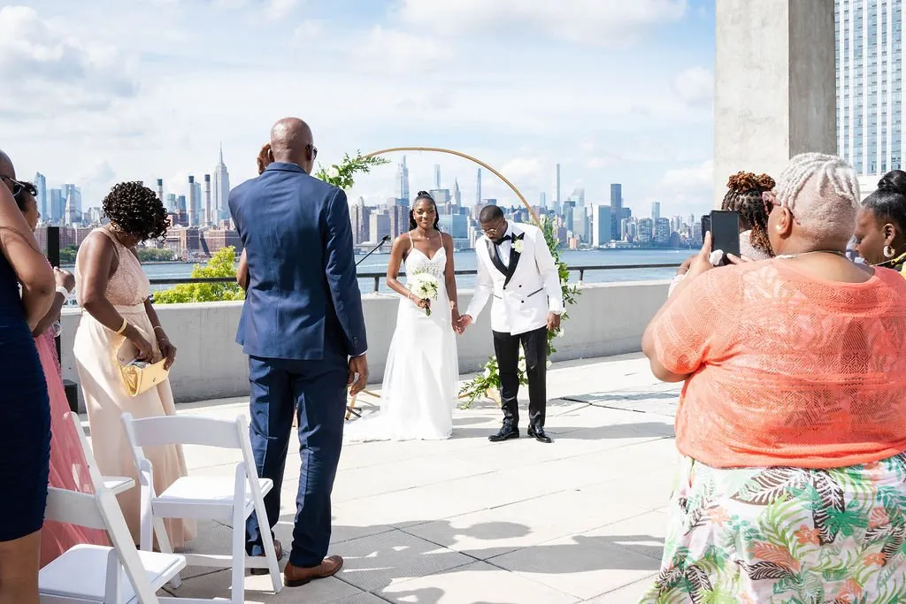 A guest stands in the aisle holding up a phone to film a rooftop wedding ceremony, the Manhattan skyline behind the couple