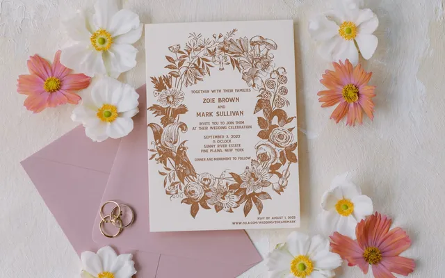 A letterpress invitation resting on a pink envelope, surrounded by cosmos flowers and a pair of rings