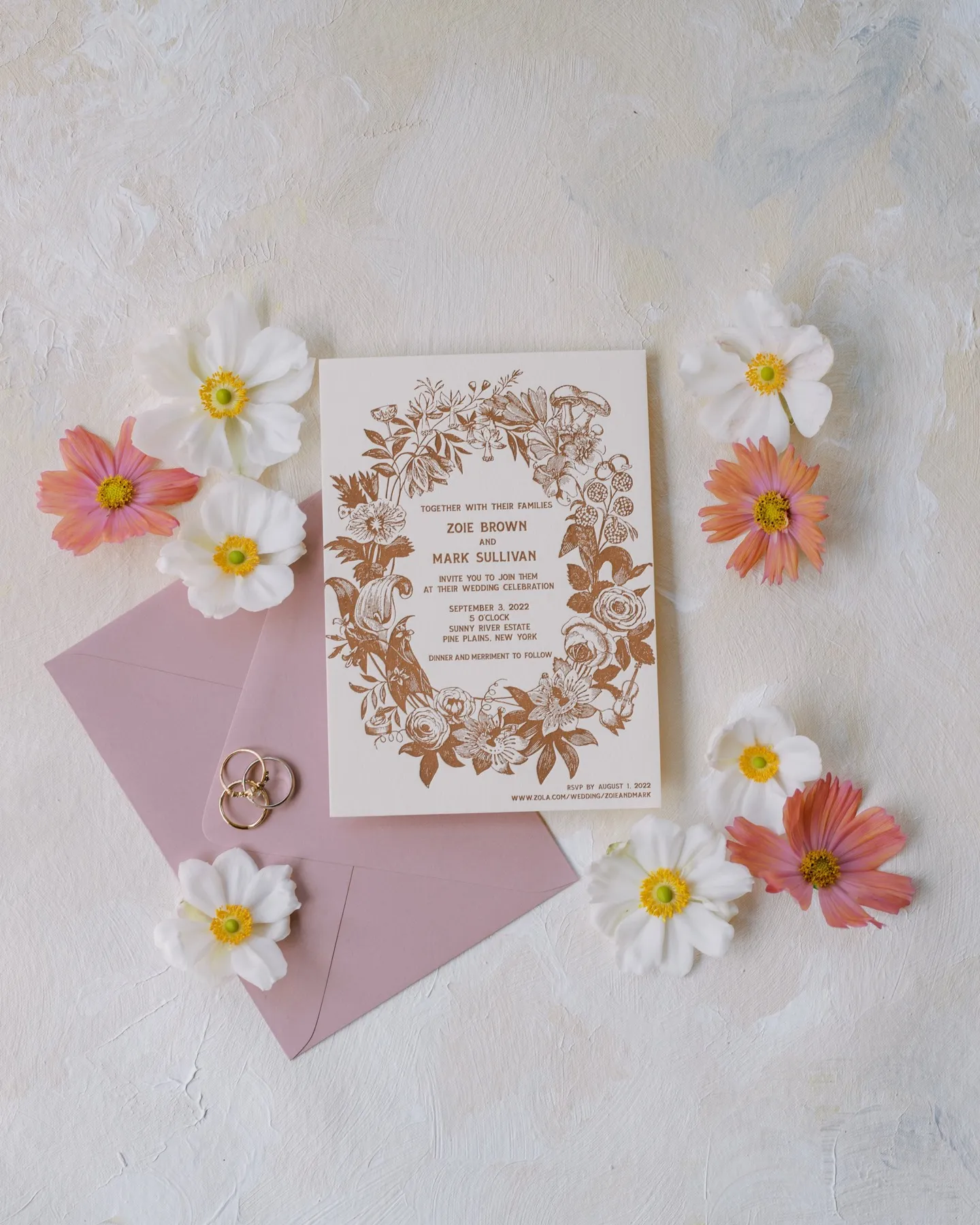 A letterpress invitation resting on a pink envelope, surrounded by cosmos flowers and a pair of rings