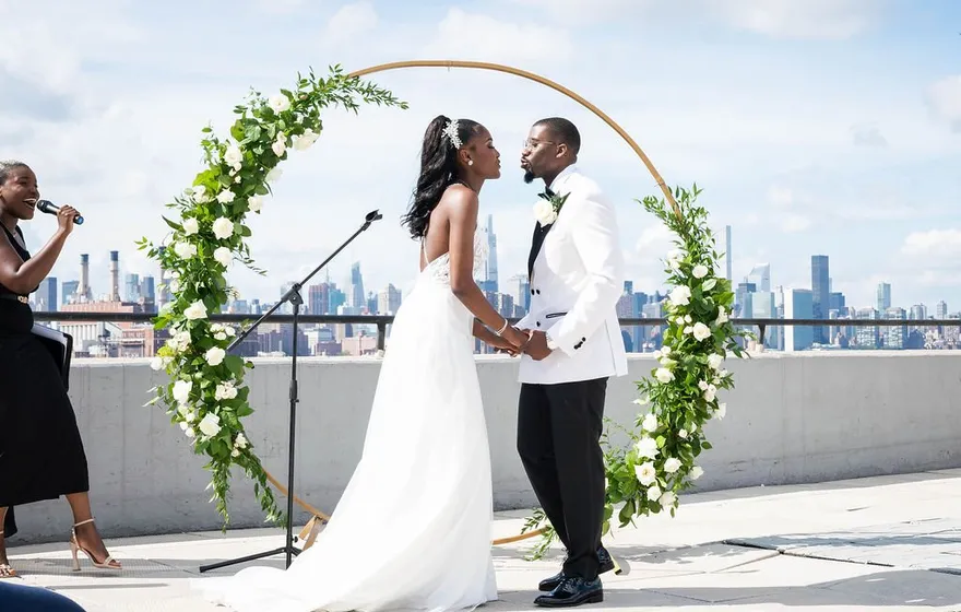 A bride and groom lean in to kiss under a round floral arch on a rooftop, the officiant at the microphone beside them and the city skyline behind