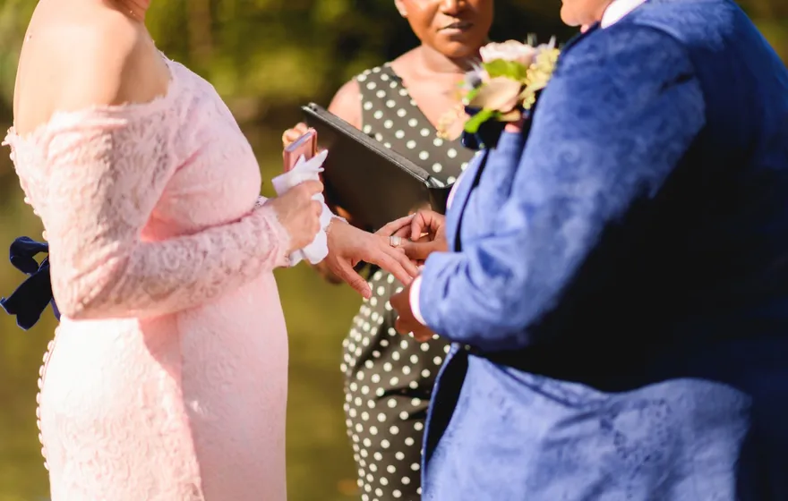 Close-up of one spouse sliding a ring onto the other's finger at a vow renewal, the officiant holding a black binder behind their hands
