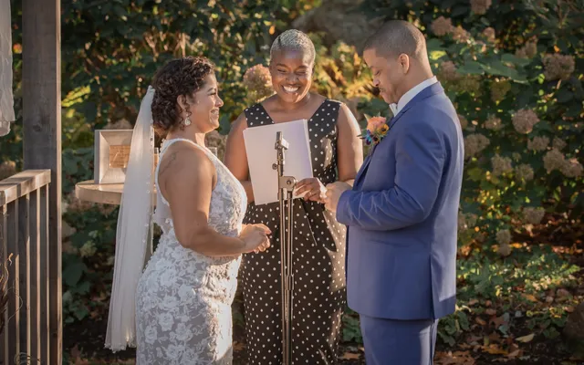 An officiant holds a printed ceremony page at a microphone and laughs with the bride and groom standing in front of her
