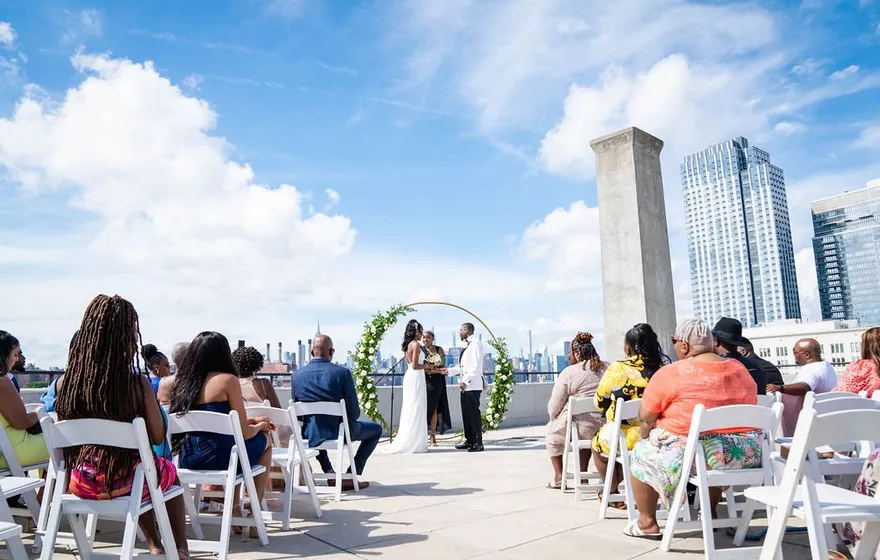 Guests seated in white folding chairs on both sides of a rooftop aisle, the couple and officiant at a floral arch at the far end with the Manhattan skyline behind
