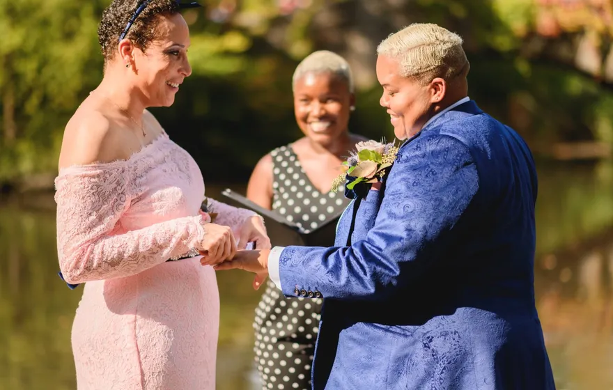 One spouse slides a ring onto the other's hand at a vow renewal while the officiant smiles behind them, binder open