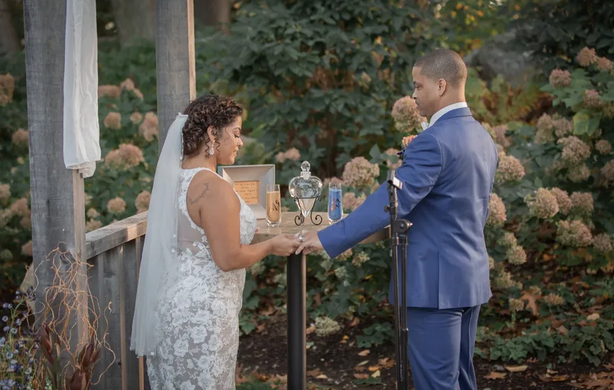A bride and groom join hands across a small table that holds a glass sand vessel during their outdoor ceremony