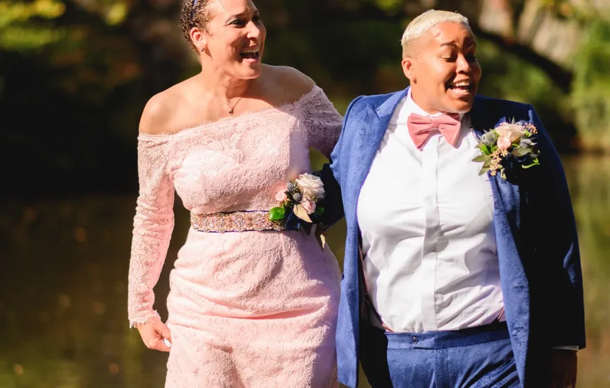 Two spouses, one in a pink lace gown and one in a blue suit and bow tie, laugh as they walk away from the water arm in arm