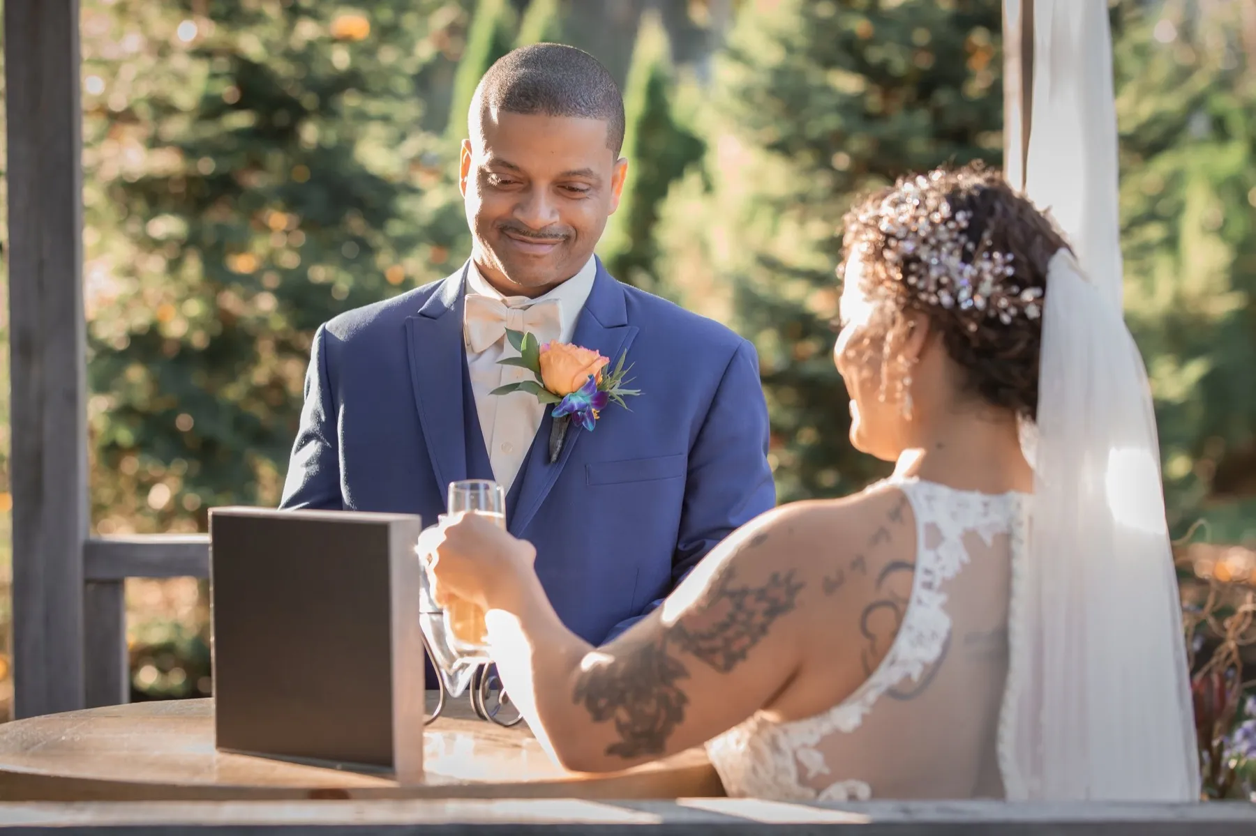 A groom and bride raise wine glasses to each other across a small table holding a wooden wine box