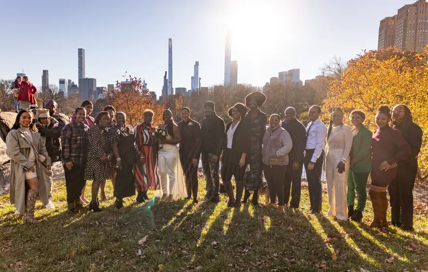 A wedding group of about two dozen people standing together on a Central Park lawn in late-afternoon sun, the Midtown skyline behind them