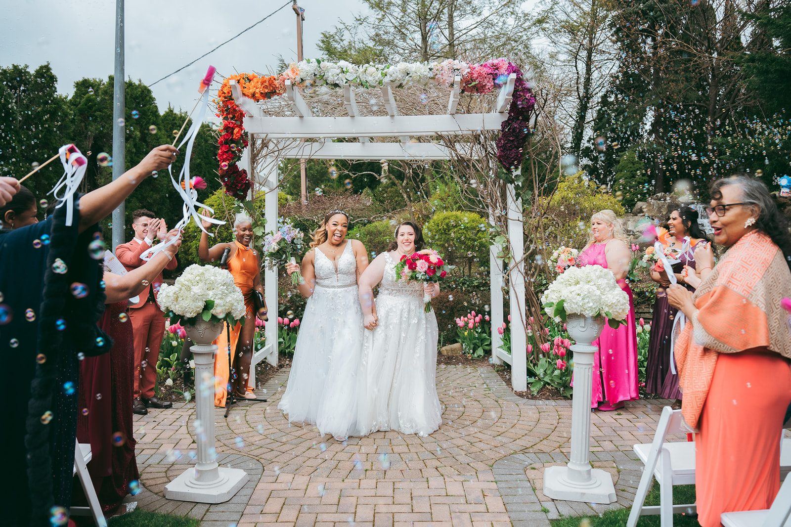 Two brides walking back up the aisle hand in hand while their guests blow bubbles