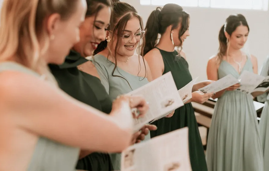 Five bridesmaids in sage and dark green gowns stand in a row in a bright room, reading open printed programs