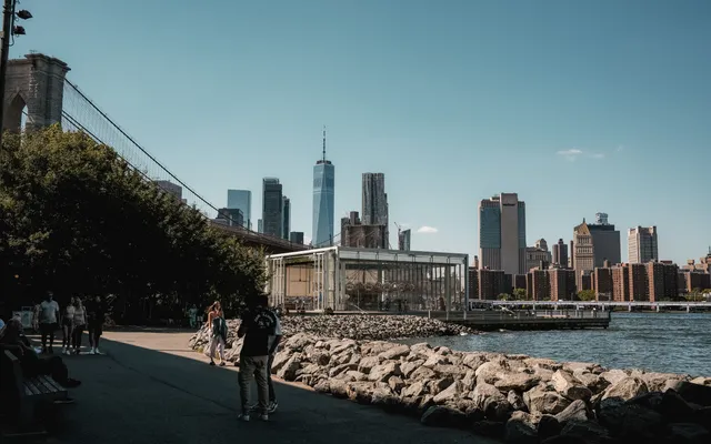 The waterfront path in Brooklyn Bridge Park beside the Brooklyn Bridge, with the glass pavilion of Jane's Carousel and the Lower Manhattan skyline across the East River
