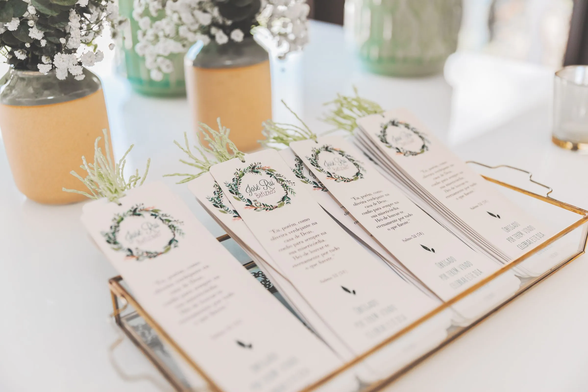 Printed wedding ceremony programs with green wreath designs fanned out in a brass tray, small jars of baby's breath behind them
