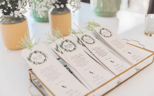 Printed wedding ceremony programs with green wreath designs fanned out in a brass tray, small jars of baby's breath behind them