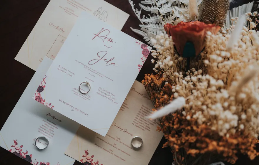 A printed invitation suite in cream and pink with a pair of gold rings resting on the cards beside a bouquet of dried flowers